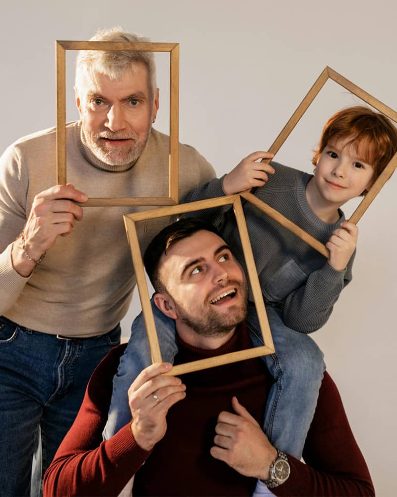 three generations of a family taking a portrait together, each member – grandpa, son, grandson – holds an empty frame over their faces