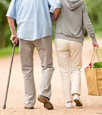 a person carrying a grocery tote in one hand holds the arm of a person walking with a cane